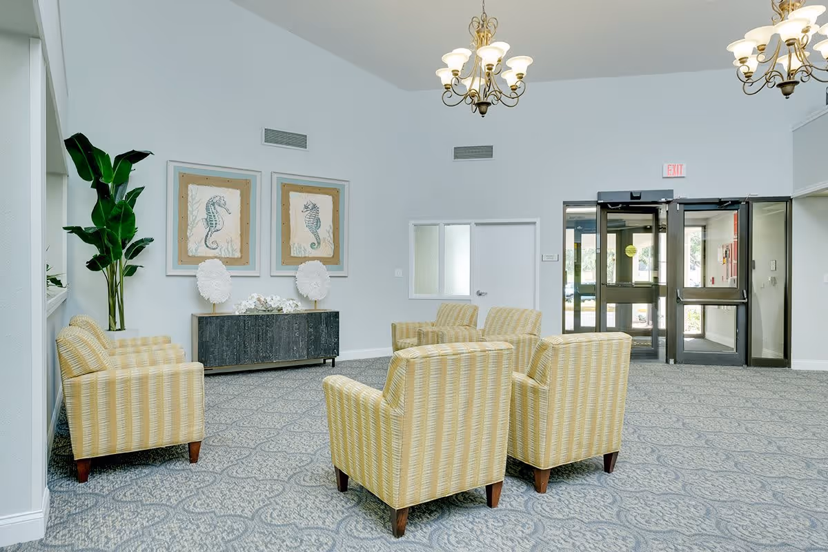 A bright and spacious seating area in a senior living facility with six yellow striped armchairs arranged in two groups. The room features a patterned carpet, two framed seahorse artworks on the wall, a large green plant, and a dark wooden console table with decorative items. There are two chandeliers hanging from the ceiling and glass double doors leading outside.
