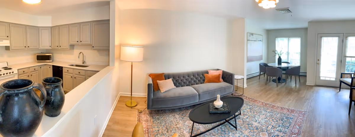 A bright and spacious living area featuring a gray tufted sofa with orange and beige pillows, a black oval coffee table with decorative items, a patterned area rug, and a floor lamp. To the left is a kitchen with beige cabinets, a white stove, microwave, and sink. In the background, there is a dining area with a round table and four chairs near large windows and a glass door leading outside.