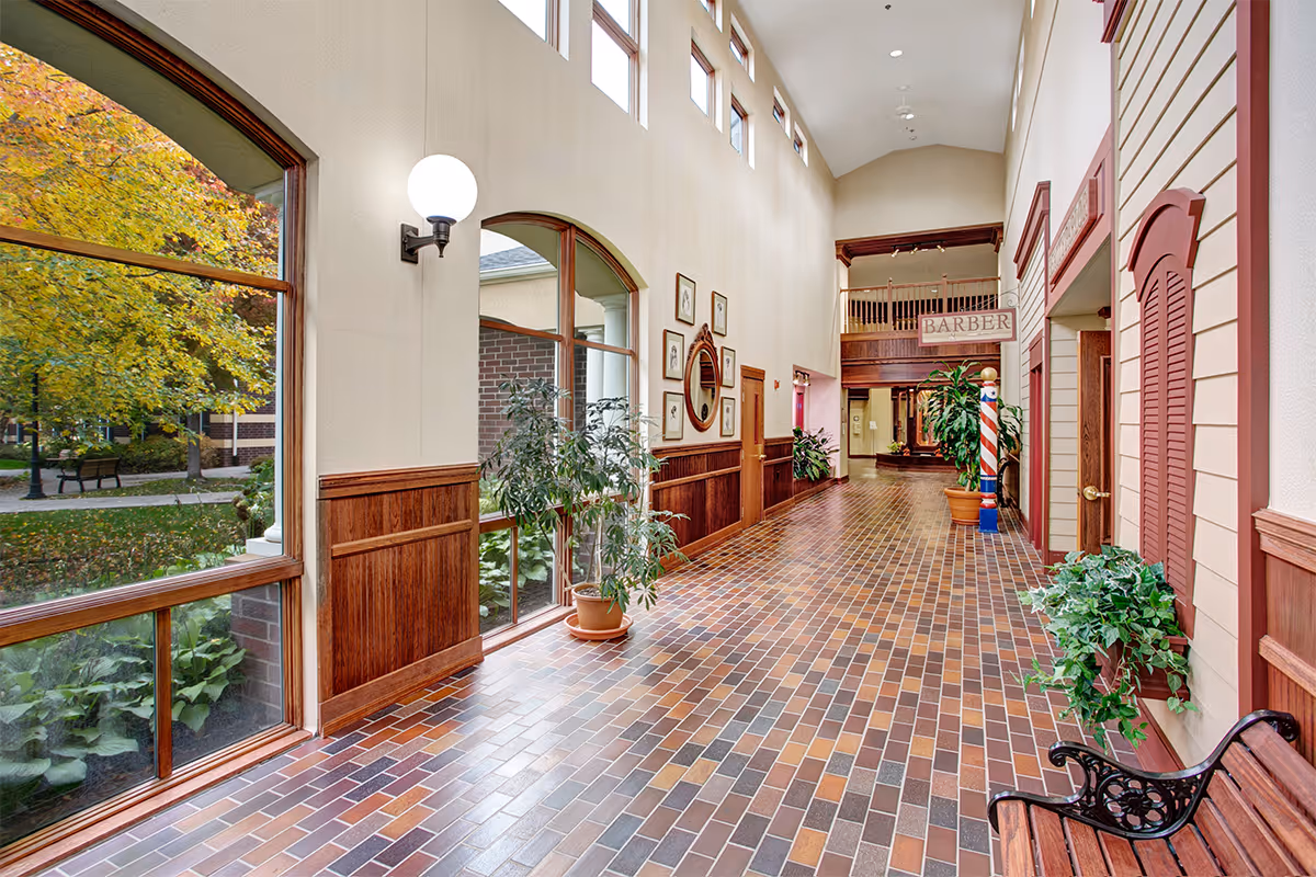 A bright hallway with large windows on the left showing autumn trees outside. The hallway has a tiled floor with various shades of brown and beige, wooden paneling on the walls, potted plants, framed pictures, and a barber pole with a sign reading 'BARBER' at the far end.