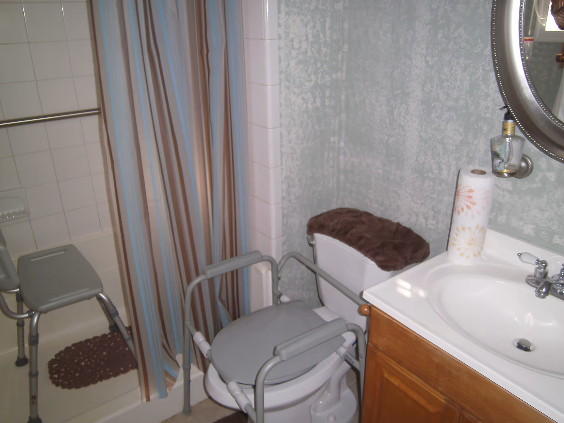 A bathroom with a shower area featuring a striped shower curtain and a shower chair. There is a toilet with a padded cover and a raised toilet seat with armrests. A sink with a wooden cabinet below and a round mirror above is also visible, along with a roll of paper towels on the sink counter.
