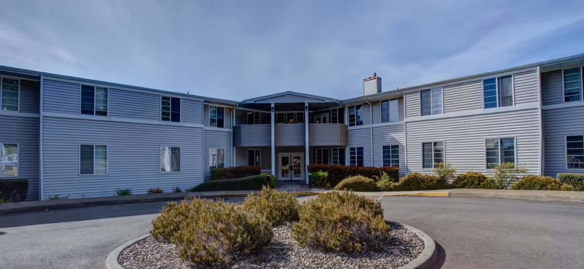 Front exterior of a two-story assisted living building with siding, a central entrance and a circular driveway with low landscaping.