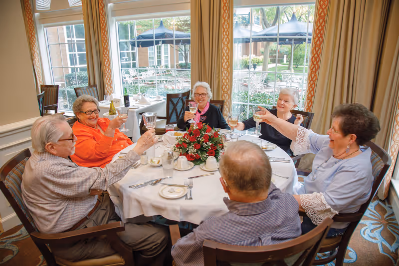 Six elderly people sitting around a round dining table in a well-lit room with large windows showing an outdoor patio. They are raising their glasses in a toast and smiling. The table is set with white tablecloth, plates, utensils, glasses, and a floral centerpiece.