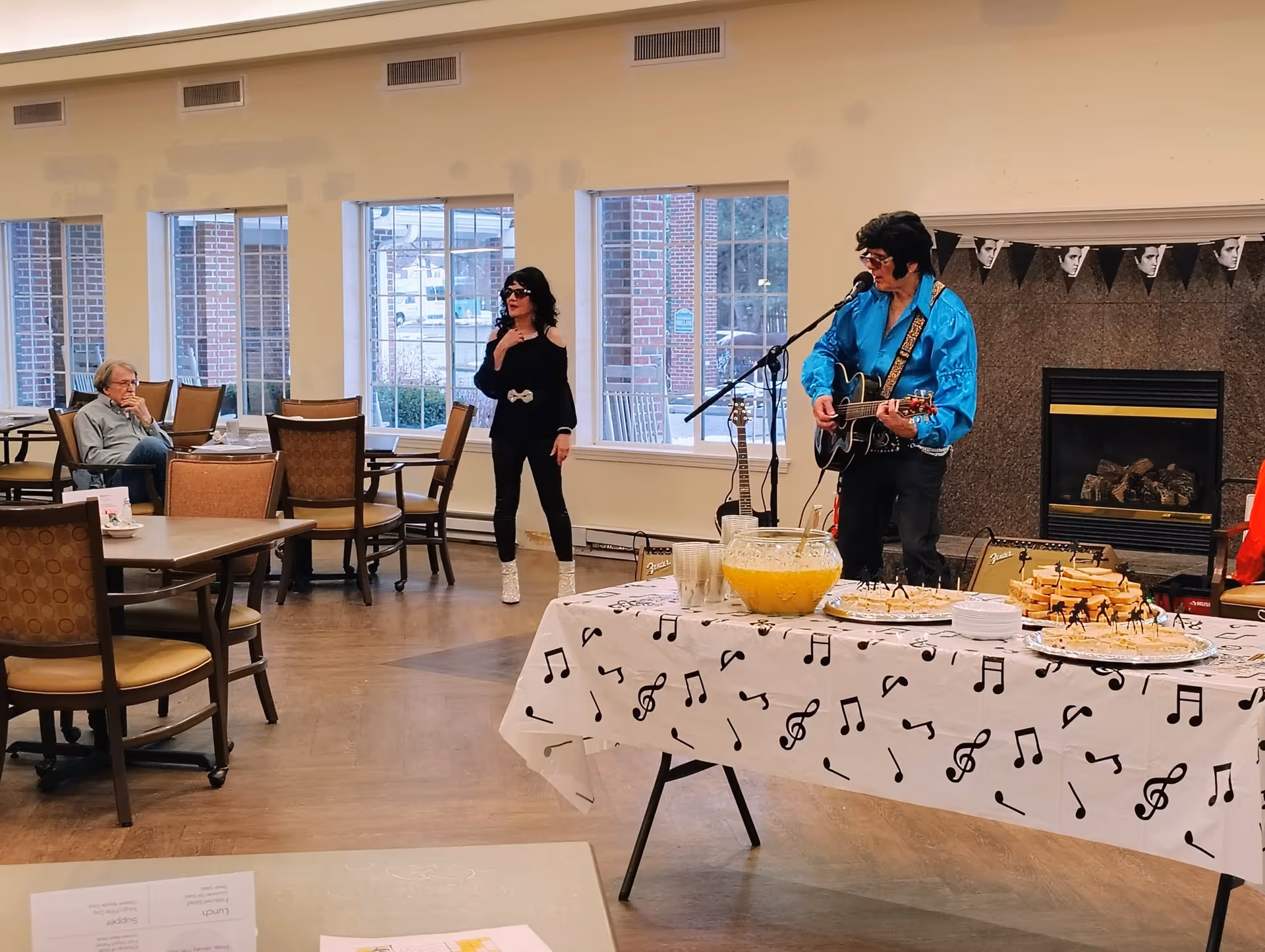 A man dressed in a blue satin shirt and black pants plays guitar and sings into a microphone, accompanied by a woman in black clothing and sunglasses standing nearby. They are performing in a room with large windows and a fireplace. In the foreground, a table covered with a white tablecloth decorated with musical notes holds plates of sandwiches and a large bowl of punch. Several empty chairs and tables are visible, with one elderly man sitting and watching the performance.