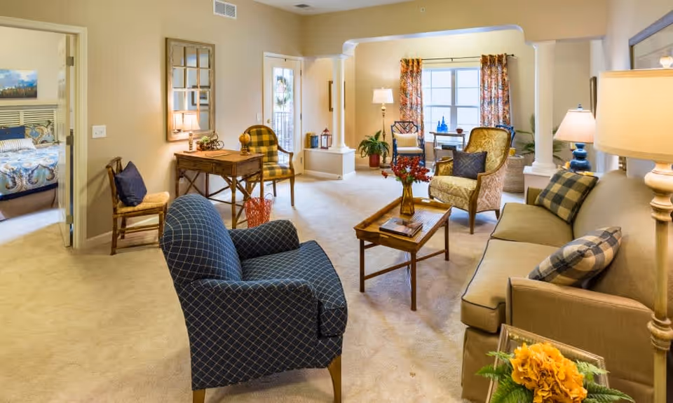 A cozy living room with a beige sofa adorned with plaid pillows, a blue patterned armchair, a wooden coffee table with a vase of flowers, and additional seating including a yellow armchair and a wooden chair. The room has beige carpeting, warm lighting from lamps, and a window with floral curtains. A bedroom with a bed and artwork is visible through an open door on the left.