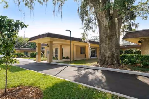 Exterior view of Camellia Gardens of Life Care with a covered entrance, driveway, and large tree.