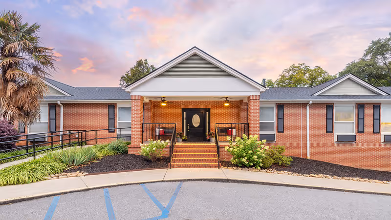 Front exterior view of Linley Park Post-Acute facility showing a single-story brick building with a covered entrance, steps leading up to a black door with sidelights, two chairs on the porch, landscaped bushes and plants, and a partly cloudy sky at sunset.