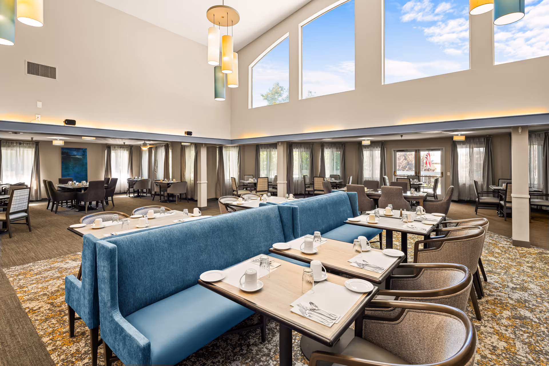 A spacious senior living community dining room with high ceilings and large windows letting in natural light. The room features multiple dining tables set with white cups, plates, and silverware. There are blue upholstered bench seats and beige cushioned chairs arranged around the tables. Modern pendant lights hang from the ceiling, and the floor is covered with a patterned carpet.