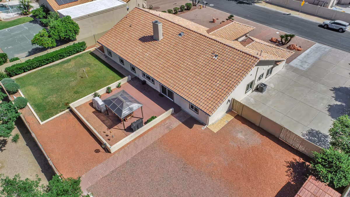 Aerial view of a single-story building with a tiled roof, surrounded by a fenced yard with a small covered patio area, green lawn, and gravel ground. The building is adjacent to a paved driveway and a street with parked cars.