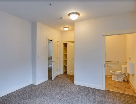 Interior view of a senior care facility room with carpeted floor, white walls, ceiling lights, an open closet with shelves, and an open door leading to a bathroom with a toilet and grab bars.