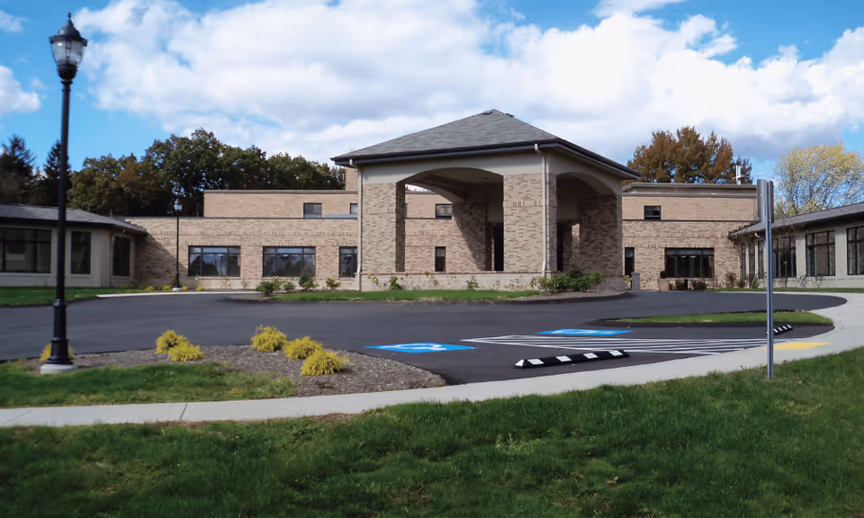 Exterior view of a single-story brick building with a covered entrance and a circular driveway with handicapped parking spaces. The building is surrounded by green grass, small shrubs, and trees under a partly cloudy blue sky.