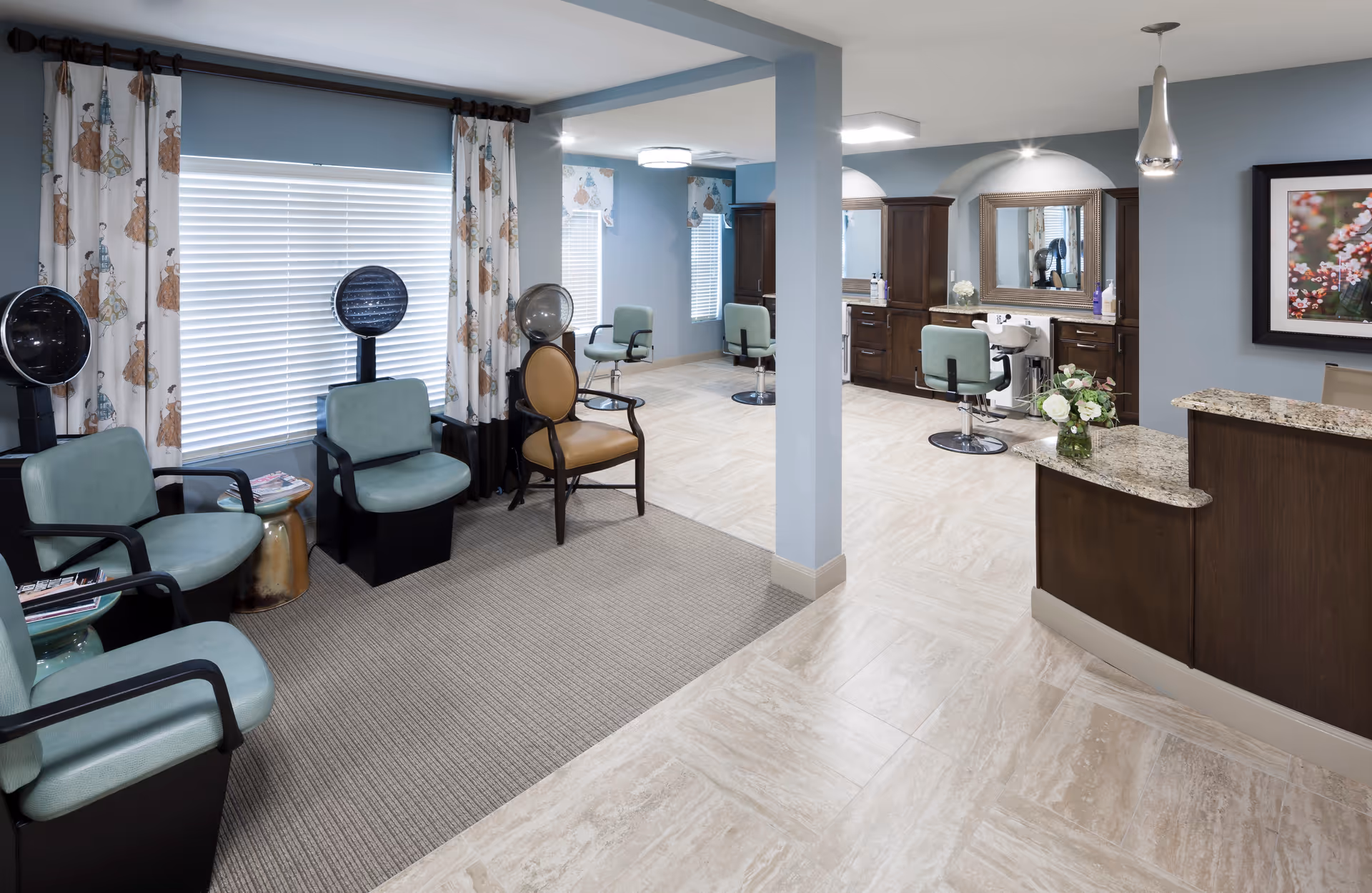 Interior view of a salon area in a senior living facility with light blue walls, multiple salon chairs, hair drying stations, a reception desk with a granite countertop, and decorative curtains on the windows.