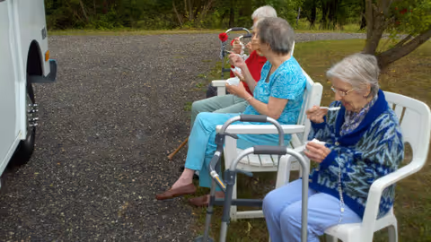 Three elderly women sitting on white plastic chairs outdoors on a gravel surface, eating ice cream. One woman has a walker in front of her. Trees and grass are visible in the background.