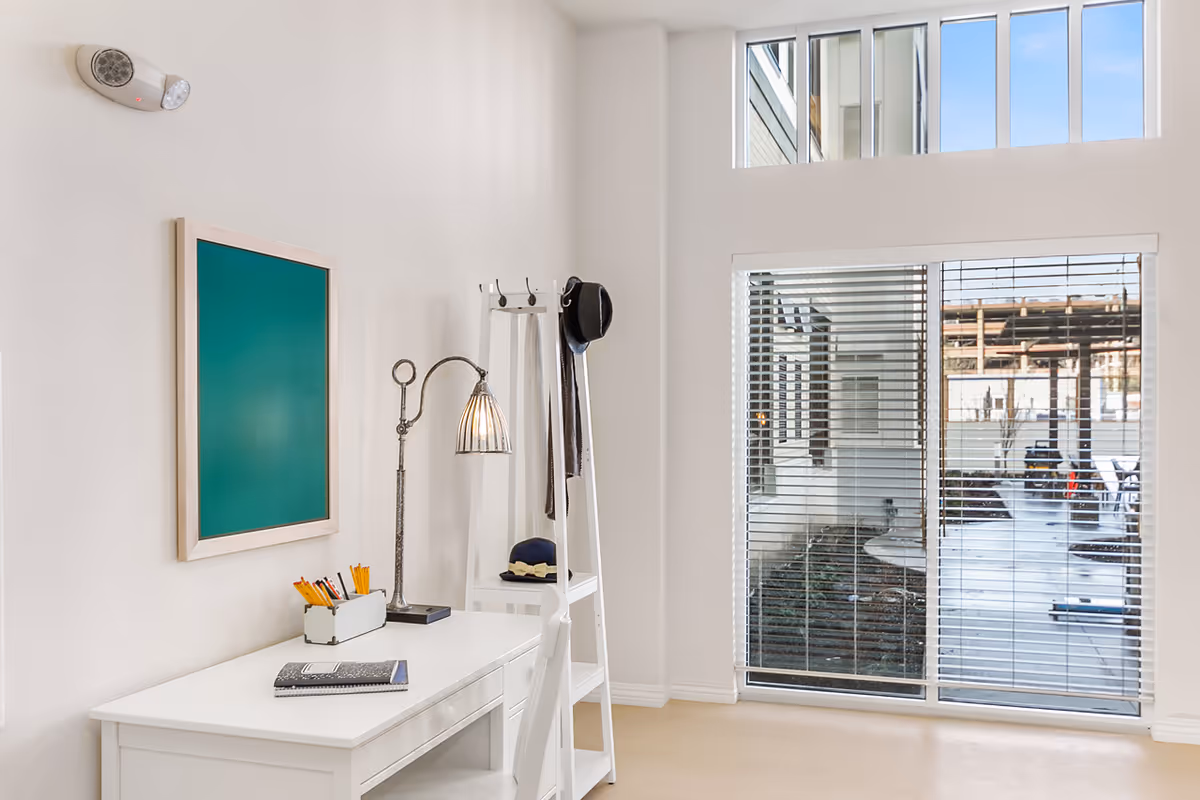 A bright room with white walls featuring a white desk with a notebook, pencils, and a vintage-style desk lamp. Next to the desk is a white ladder shelf with a hat and scarf hanging on it. A large window with blinds looks out onto an outdoor patio area with seating and a pergola.