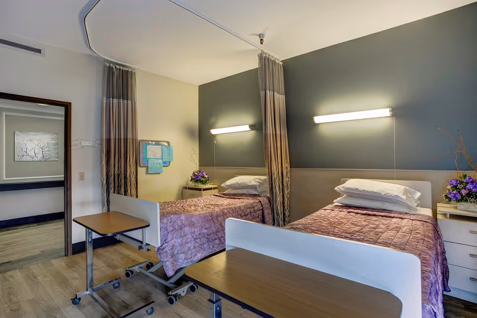 A shared bedroom in a senior living facility with two single beds, privacy curtains, and bedside tables.