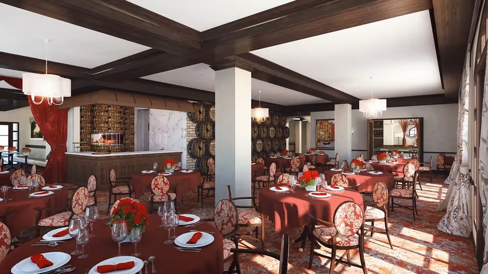 A spacious dining room with round tables covered in red tablecloths, each set with white plates, red napkins, wine glasses, and silverware. The chairs have a floral upholstery pattern. The room features wooden ceiling beams, large windows with patterned curtains allowing natural light, and a decorative fireplace with a brick surround. Wine barrels are stacked against one wall, and there are chandeliers hanging from the ceiling.