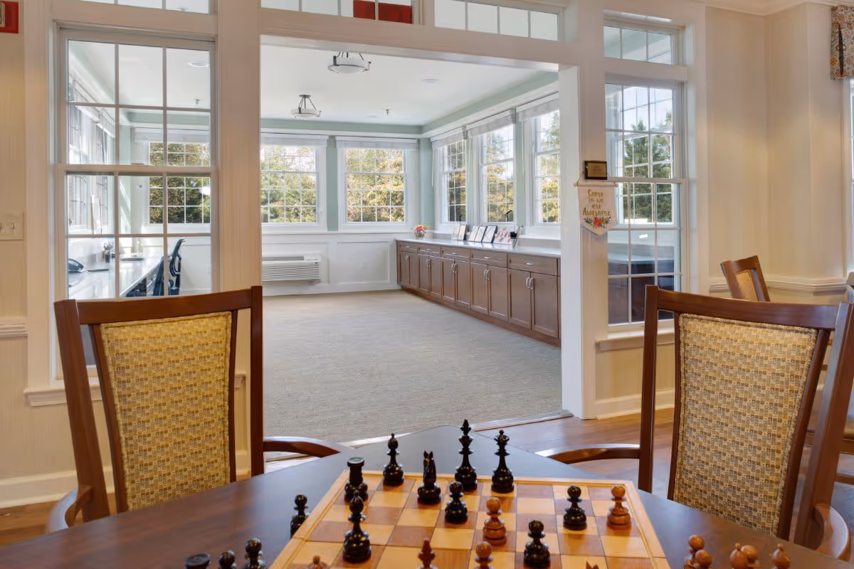A bright and spacious common area with large windows letting in natural light. In the foreground, there is a wooden table with a chessboard and chess pieces set up, surrounded by cushioned wooden chairs. Beyond the table, there is an open room with carpeted flooring, wooden cabinets along the wall, and more windows showing greenery outside.
