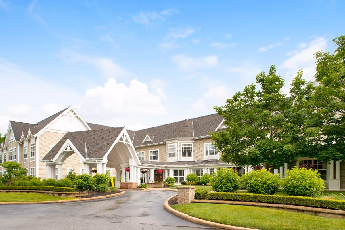 Exterior view of Sunrise of Cuyahoga Falls senior living facility showing a large, two-story building with beige siding, white trim, and a covered entrance. The driveway curves around landscaped greenery including bushes, trees, and manicured lawns under a partly cloudy blue sky.