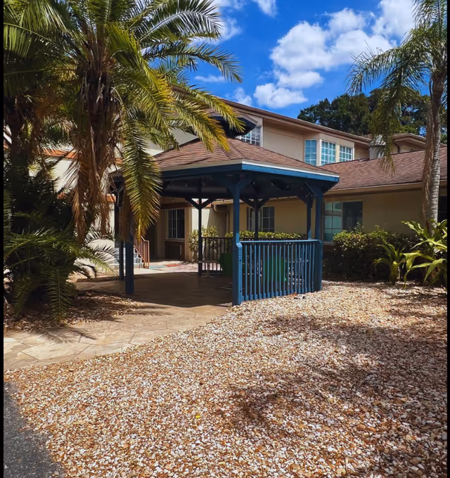 Outdoor area of a senior living facility featuring a blue wooden gazebo surrounded by palm trees and other greenery, with a building in the background under a partly cloudy blue sky.