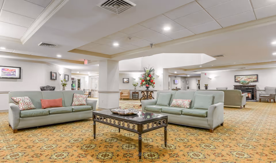 Spacious senior living community lounge area with multiple light green sofas adorned with patterned and solid cushions, a dark wooden coffee table with decorative items, floral arrangements on side tables, patterned carpet flooring, and soft lighting from ceiling fixtures and wall sconces.
