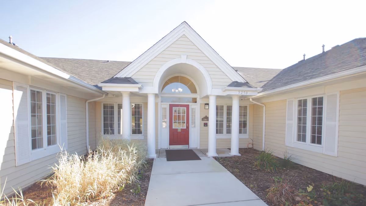 Front entrance of a single-story beige building with white columns, a red door, and a concrete walkway.