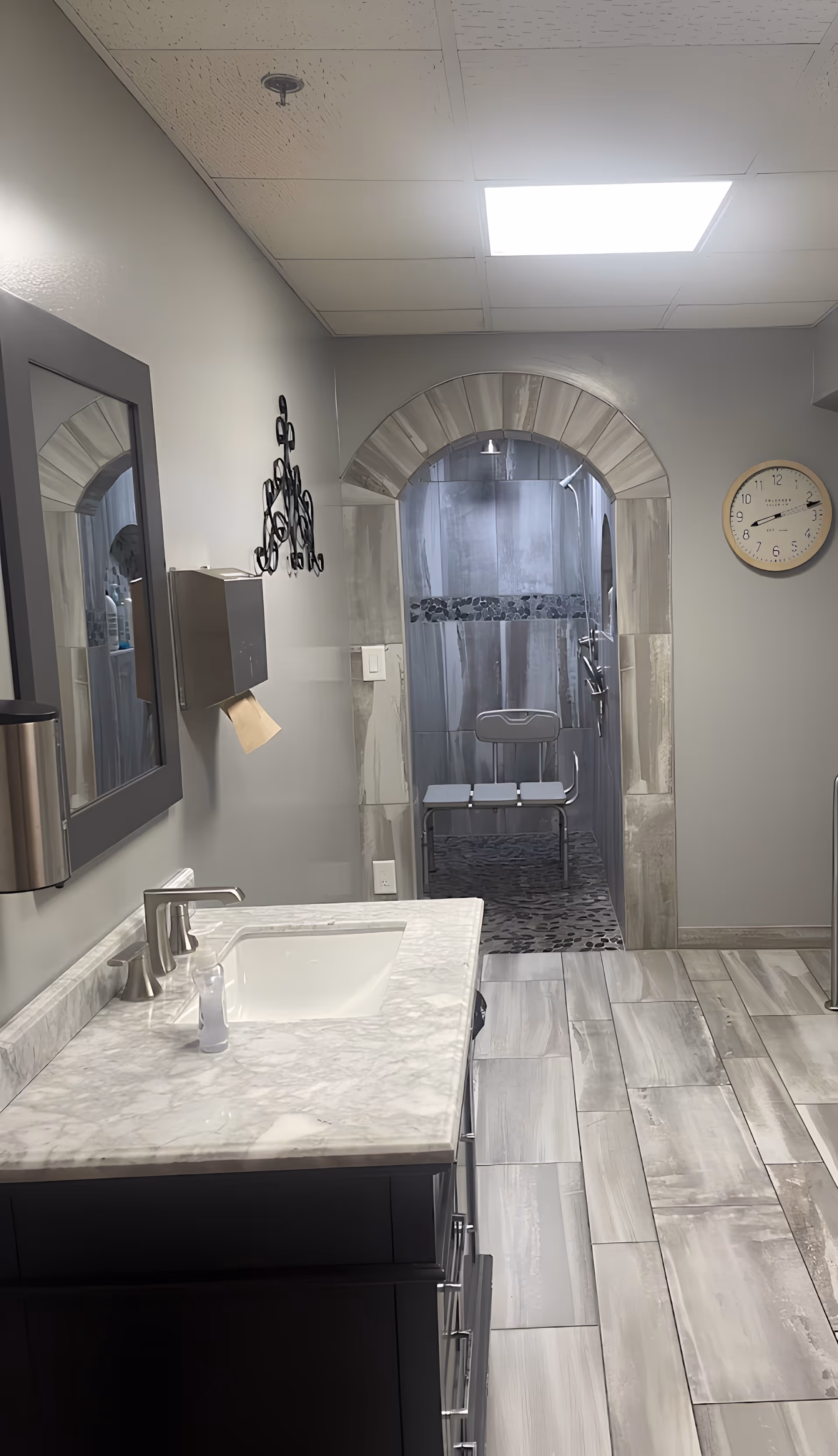 A modern bathroom with a marble countertop sink, a large mirror, and a wall-mounted paper towel dispenser. The room features a tiled floor and an arched entrance leading to a walk-in shower area with a shower chair and pebble stone flooring. A clock is mounted on the wall to the right.
