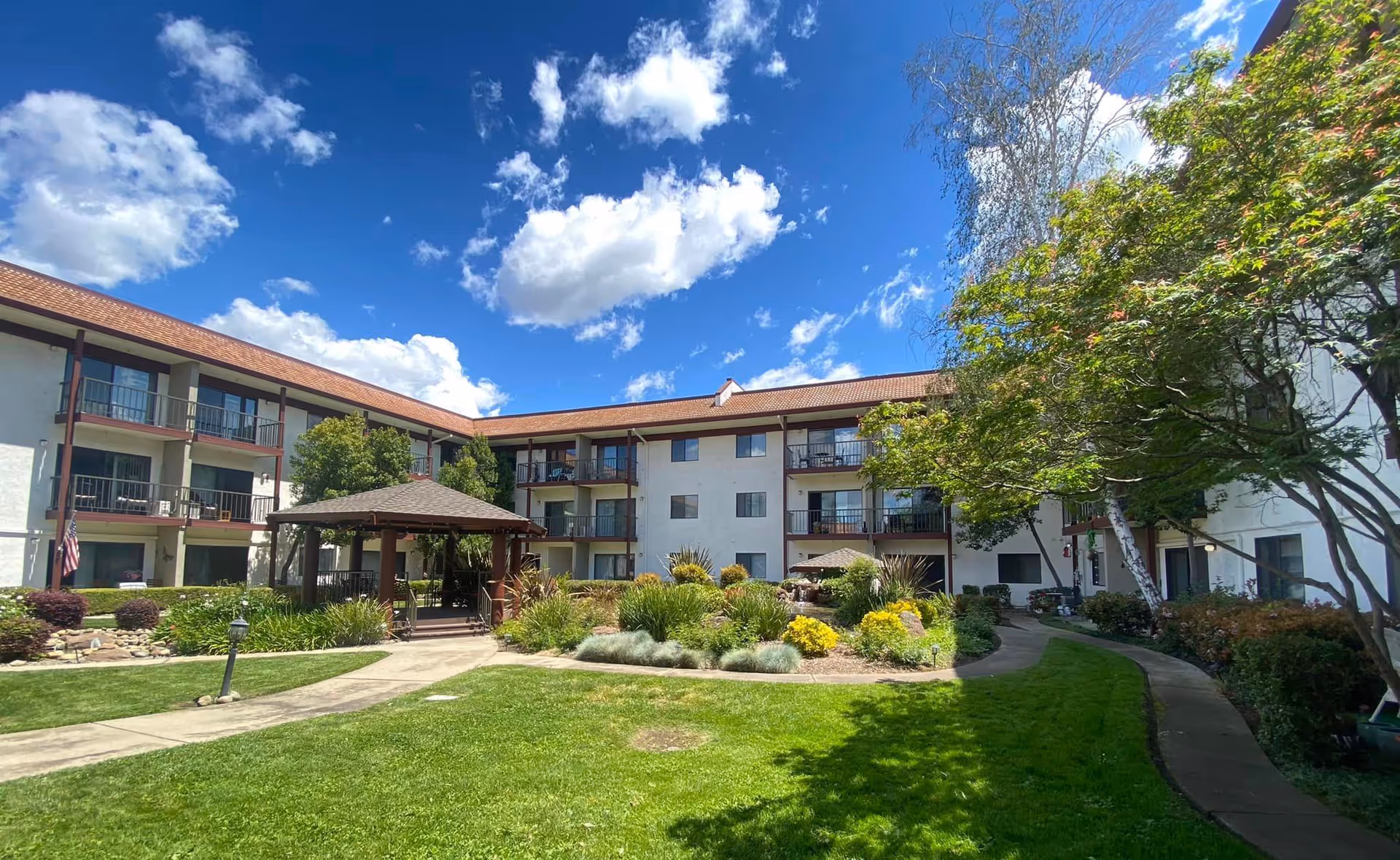 Outdoor courtyard area of Roseville Commons Senior Living with a well-maintained lawn, a gazebo, various shrubs and trees, and a three-story building with balconies surrounding the courtyard under a partly cloudy blue sky.