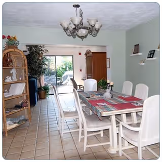 A dining room with a rectangular table covered with a colorful tablecloth and surrounded by six white cushioned chairs. There is a chandelier hanging from the ceiling above the table. To the left, there is a wooden shelving unit with decorative items and a large potted plant near a sliding glass door that leads to an outdoor patio area with chairs and greenery visible outside.