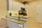 A modern kitchen area with granite countertops, white cabinets, a stainless steel microwave and stove, a small orchid plant, and a bottle of wine with two glasses on the counter.