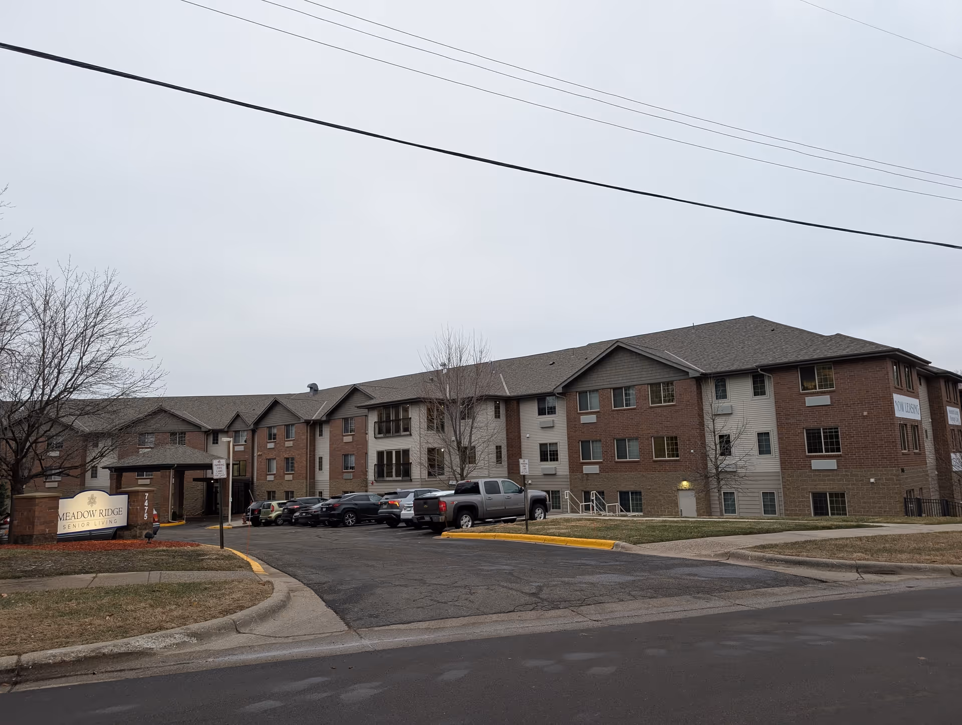 Exterior view of Meadow Ridge Senior Living building with a parking lot in front. The building is three stories tall with a combination of brick and siding exterior. There is a sign near the entrance that reads 'Meadow Ridge Senior Living'. Several cars are parked in the lot and leafless trees are visible around the property under an overcast sky.