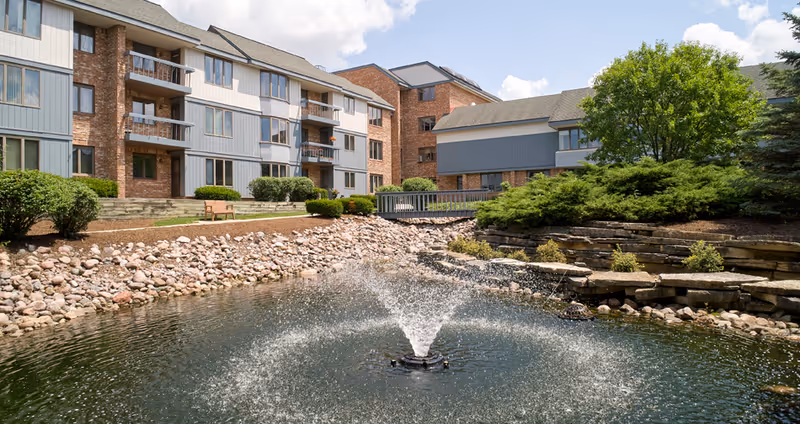 Outdoor view of a senior living facility with a pond featuring a central water fountain, surrounded by rocks and landscaped greenery. The building in the background has multiple floors with balconies and large windows under a partly cloudy sky.
