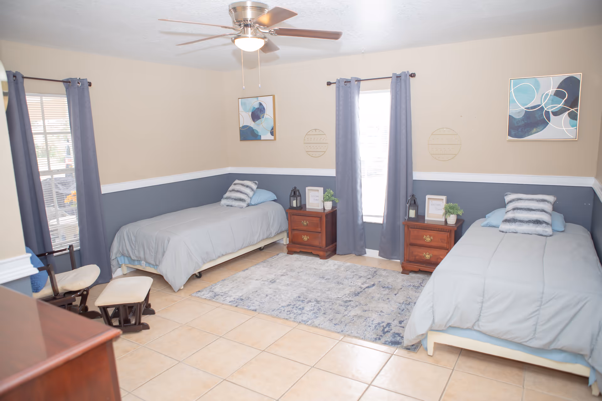 A bedroom with two single beds, each with gray bedding and a striped pillow. Between the beds are two wooden nightstands with decorative items including lanterns, a small plant, and framed signs. The room has beige and blue walls with white trim, two windows with gray curtains, a ceiling fan with a light, a blue patterned rug on the tiled floor, and a wooden rocking chair with a matching footrest.
