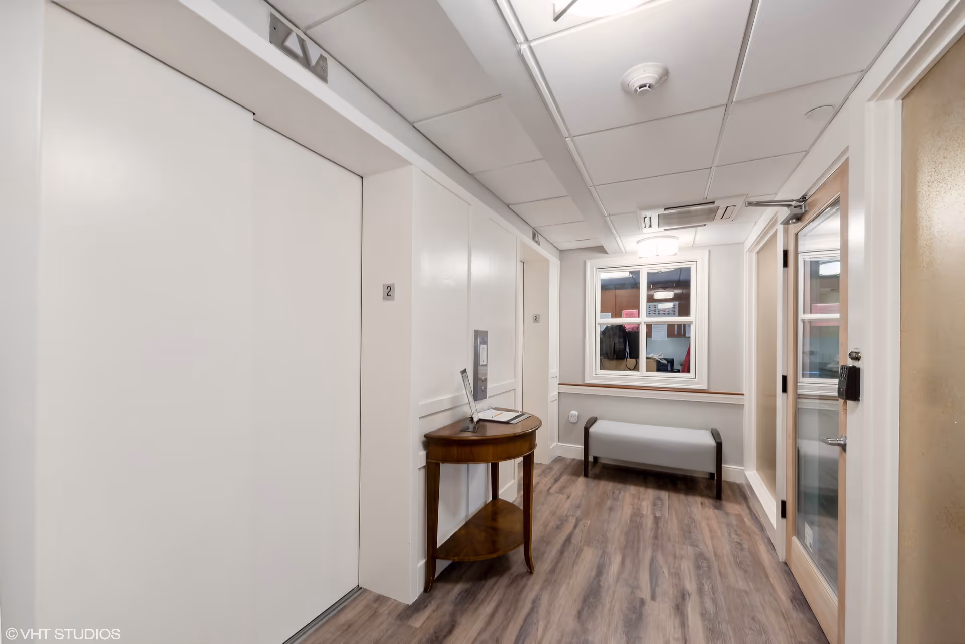 A clean, well-lit hallway with two white elevator doors labeled with the number 2, a small wooden table with a sign and papers on it, a window showing an interior room, a gray bench beneath the window, and a glass door with a keypad lock on the right side. The floor is wood-patterned and the ceiling has recessed lighting and a smoke detector.