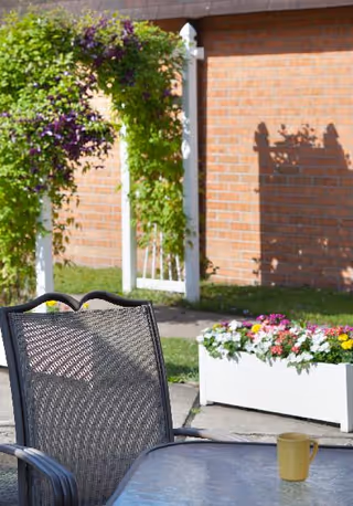 Outdoor patio area with a metal mesh chair and a glass-top table with a yellow mug on it. In the background, there is a white trellis with green and purple flowering vines, a white rectangular planter box filled with colorful flowers, and a brick wall.