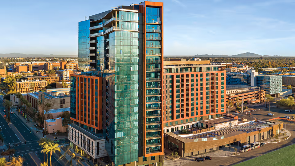 Aerial view of a modern multi-story building with glass and orange architectural accents, surrounded by city streets and other buildings under a clear sky.