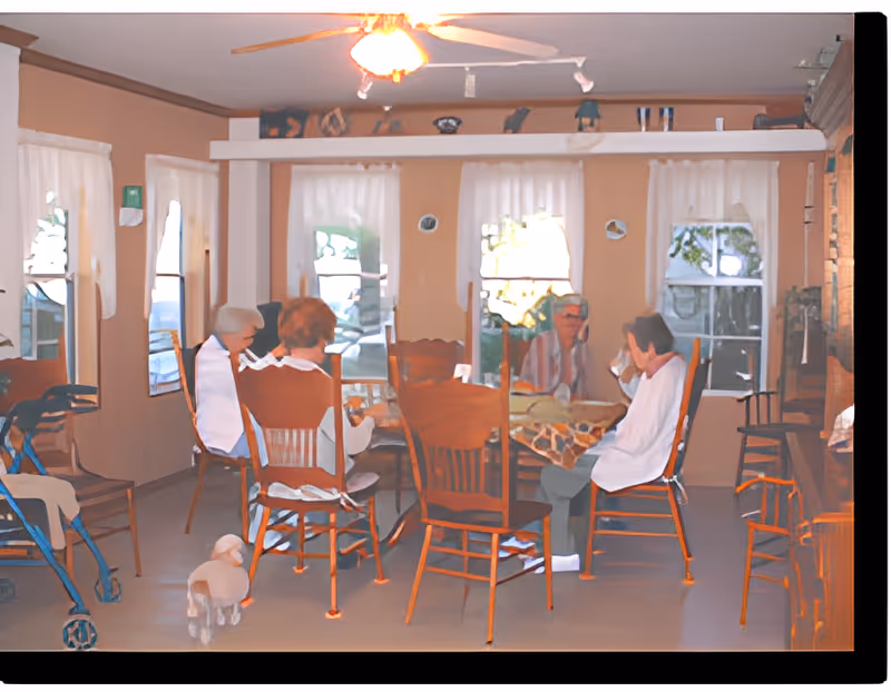 A group of elderly people sitting around a wooden dining table in a room with beige walls and several windows with white curtains. There is a ceiling fan with a light above the table, and a small dog is on the floor near the chairs. A walker is visible on the left side of the image.