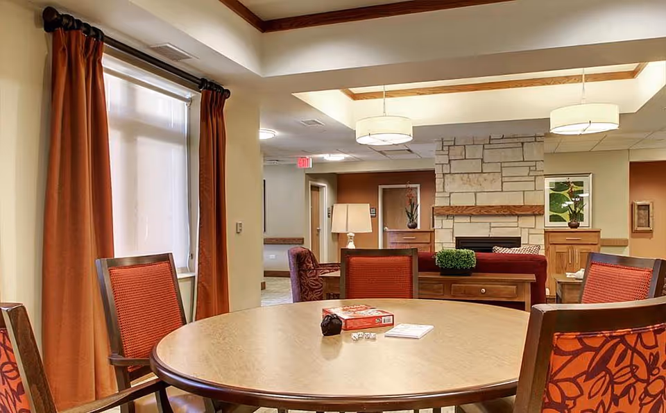 Communal dining area with a round table and red-upholstered chairs, a stone fireplace and warm overhead lighting.