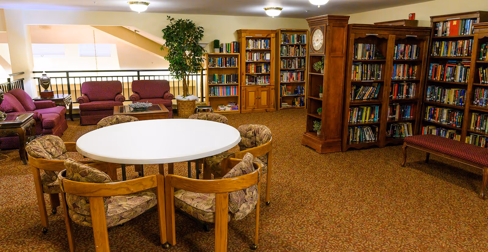 A cozy library room with multiple wooden bookshelves filled with books, a large wooden grandfather clock, a round white table surrounded by six cushioned chairs, and a seating area with three maroon upholstered armchairs and a small table. The room has warm lighting and carpeted flooring with a patterned design.