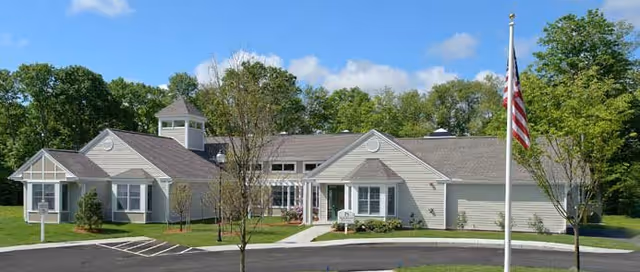 Exterior view of a single-story senior living facility building named White Oak Cottages, featuring beige siding, multiple peaked roofs, a small cupola, and an American flag on a flagpole in the front lawn. The building is surrounded by green grass, small trees, and a paved driveway with parking spaces.