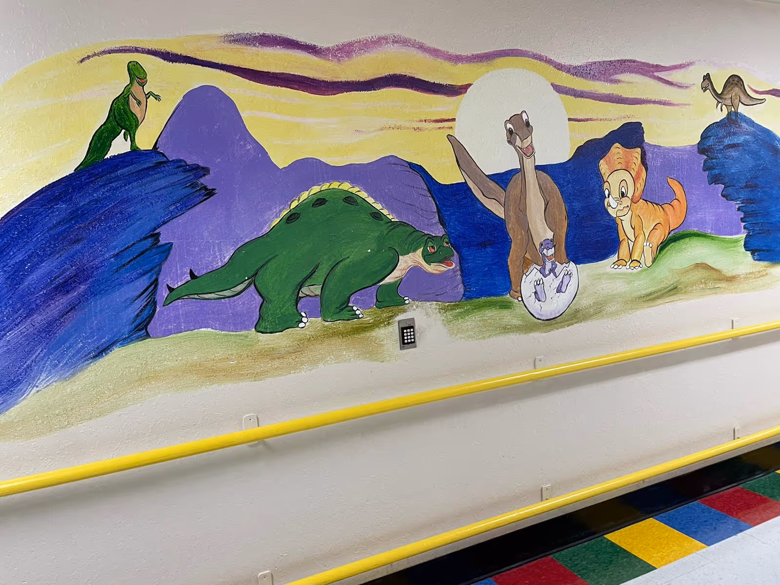 A colorful mural painted on a wall depicting cartoon dinosaurs in a prehistoric landscape with purple mountains and a yellow sky. The scene includes a green dinosaur on a rock, a large green dinosaur walking, a dinosaur hatching from an egg, and two other dinosaurs standing on the ground and rocks. The wall has a yellow handrail and a multicolored tiled floor below.