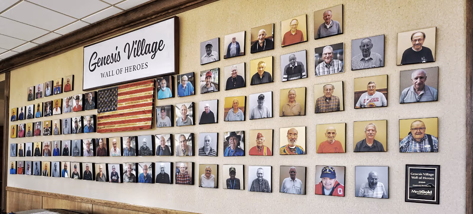 A wall display titled 'Genesis Village Wall of Heroes' featuring numerous framed photographs of elderly individuals arranged in rows around a wooden American flag. The display honors residents or veterans and is mounted on a beige wall above a wooden bench.