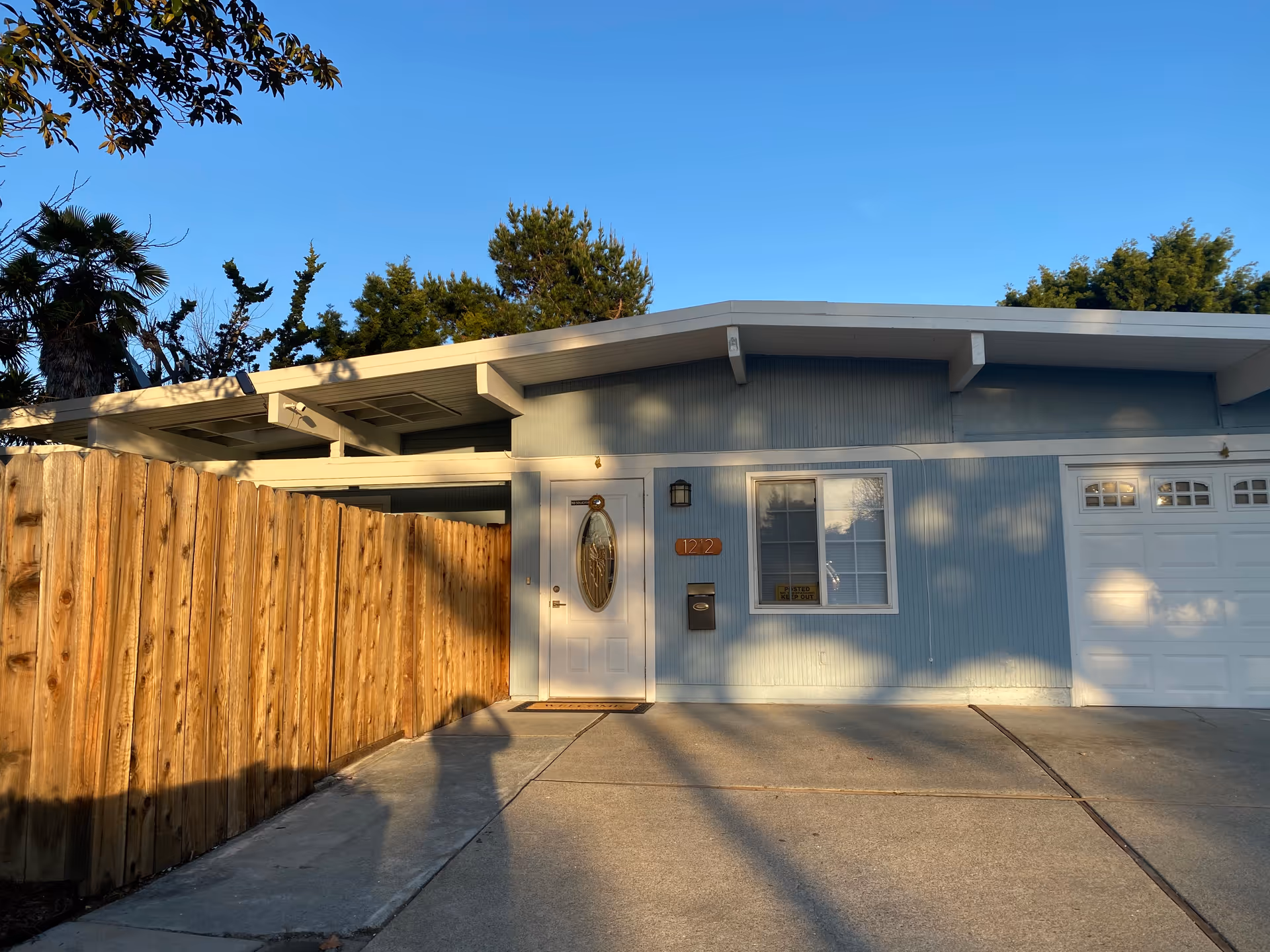 Front exterior of a light-blue single-story house with a wooden fence, driveway, front door, and attached garage.