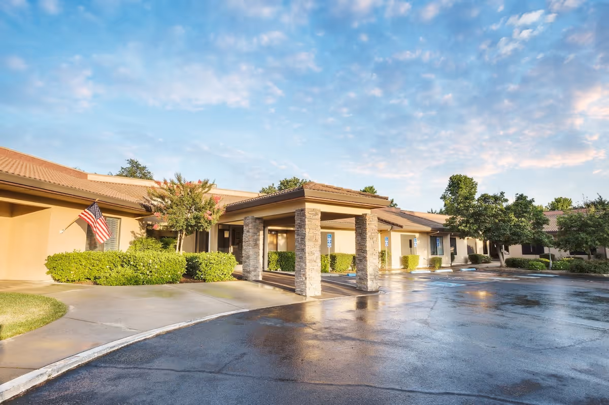 Exterior view of Copper Ridge Care Center showing a single-story building with a covered entrance supported by stone pillars, surrounded by greenery and an American flag on the left side. The sky is partly cloudy and the parking area is wet, reflecting the building and trees.