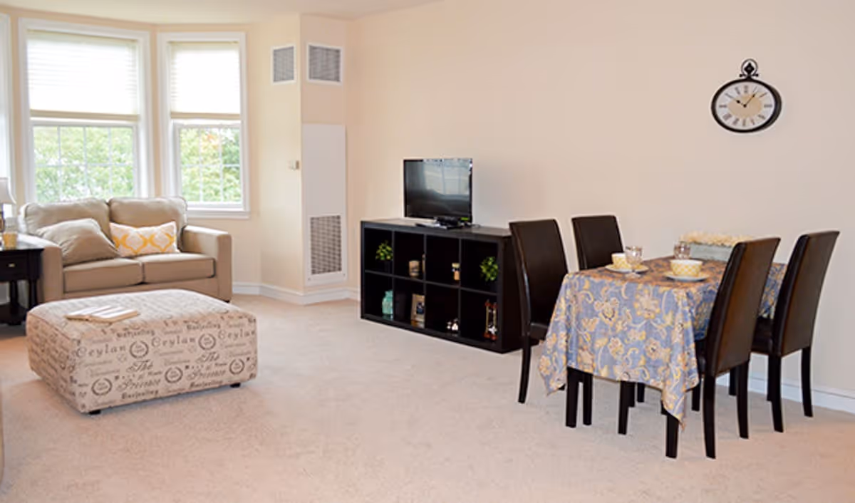 A cozy living and dining area with a beige loveseat, a patterned ottoman, a black shelving unit with a TV on top, and a dining table covered with a blue and gold patterned tablecloth surrounded by four dark chairs. A wall clock is mounted on the beige wall above the dining table, and three windows with white blinds let in natural light.