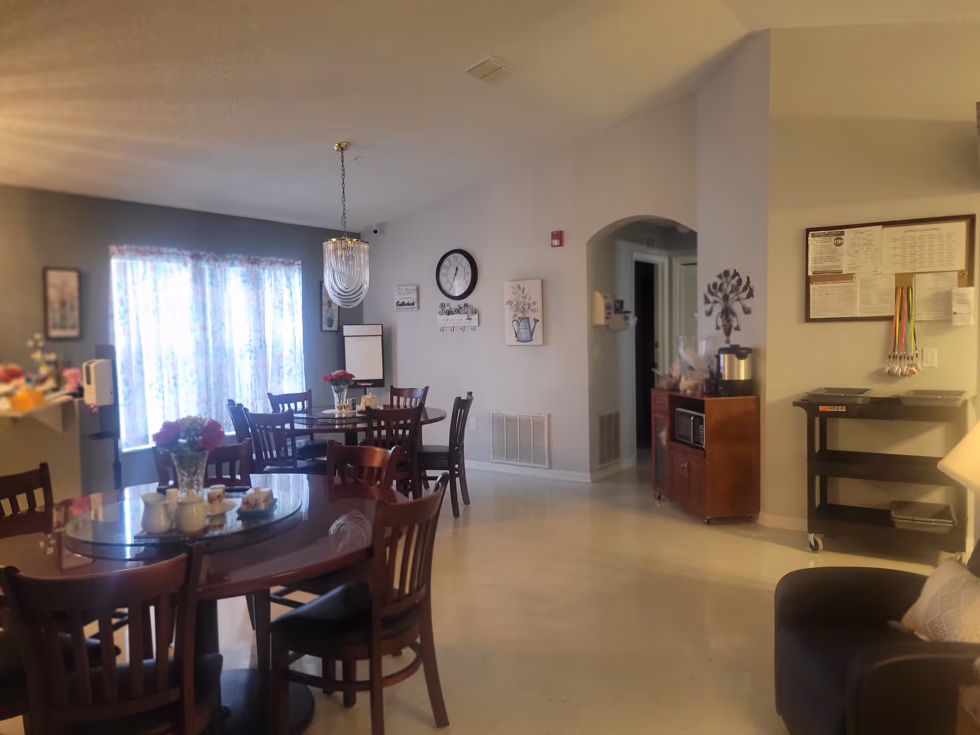 Communal dining area with round tables and chairs, a chandelier, and a small service cart against the wall.