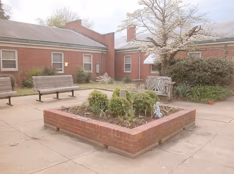 Outdoor courtyard area with brick raised flower bed containing green plants and flowers, surrounded by concrete pavement. Several wooden benches are placed around the courtyard. The background shows a red brick building with windows and a tree with light-colored blossoms.
