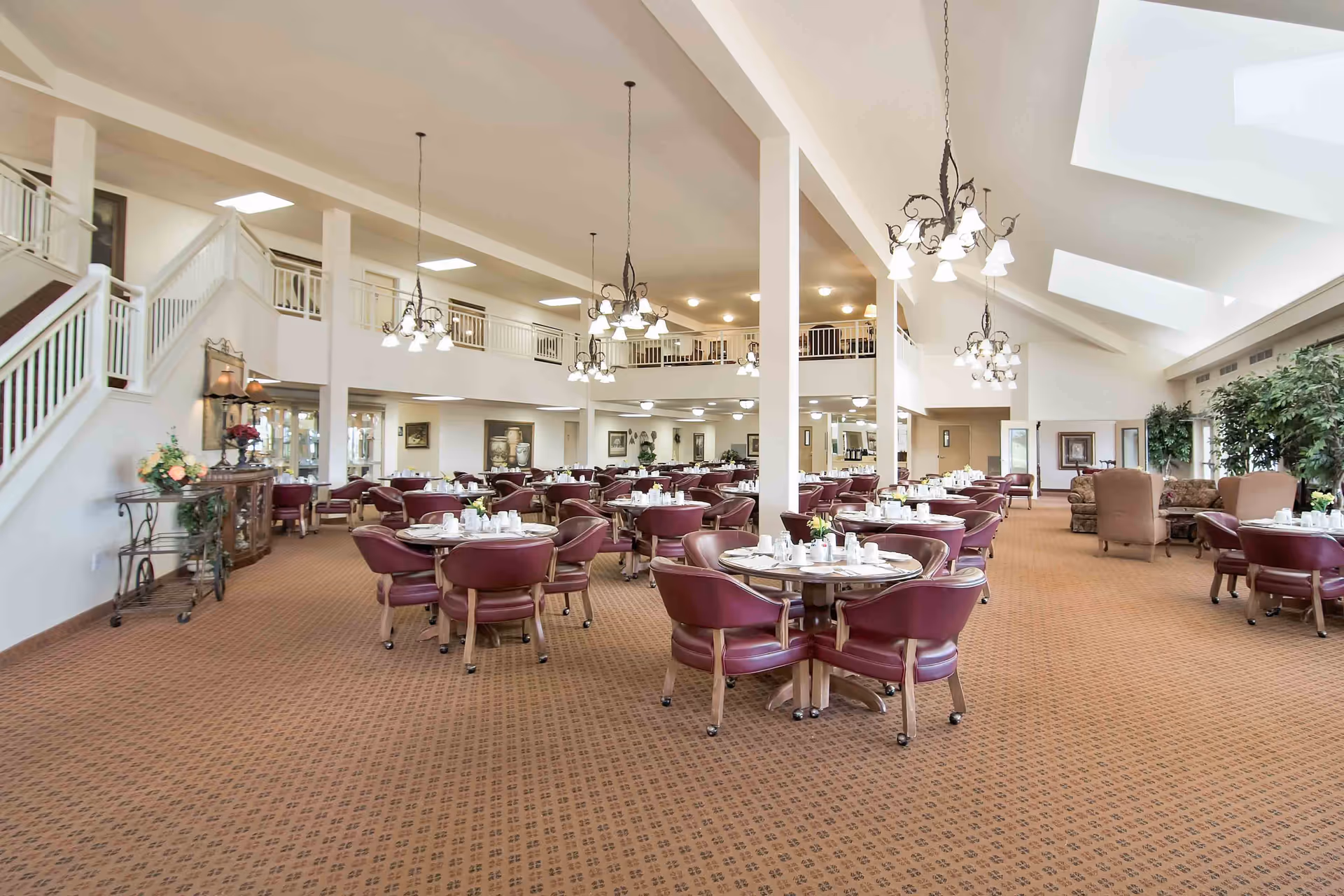 Spacious two-story dining room with round tables set for meals, maroon chairs, chandeliers, and an upper balcony walkway.