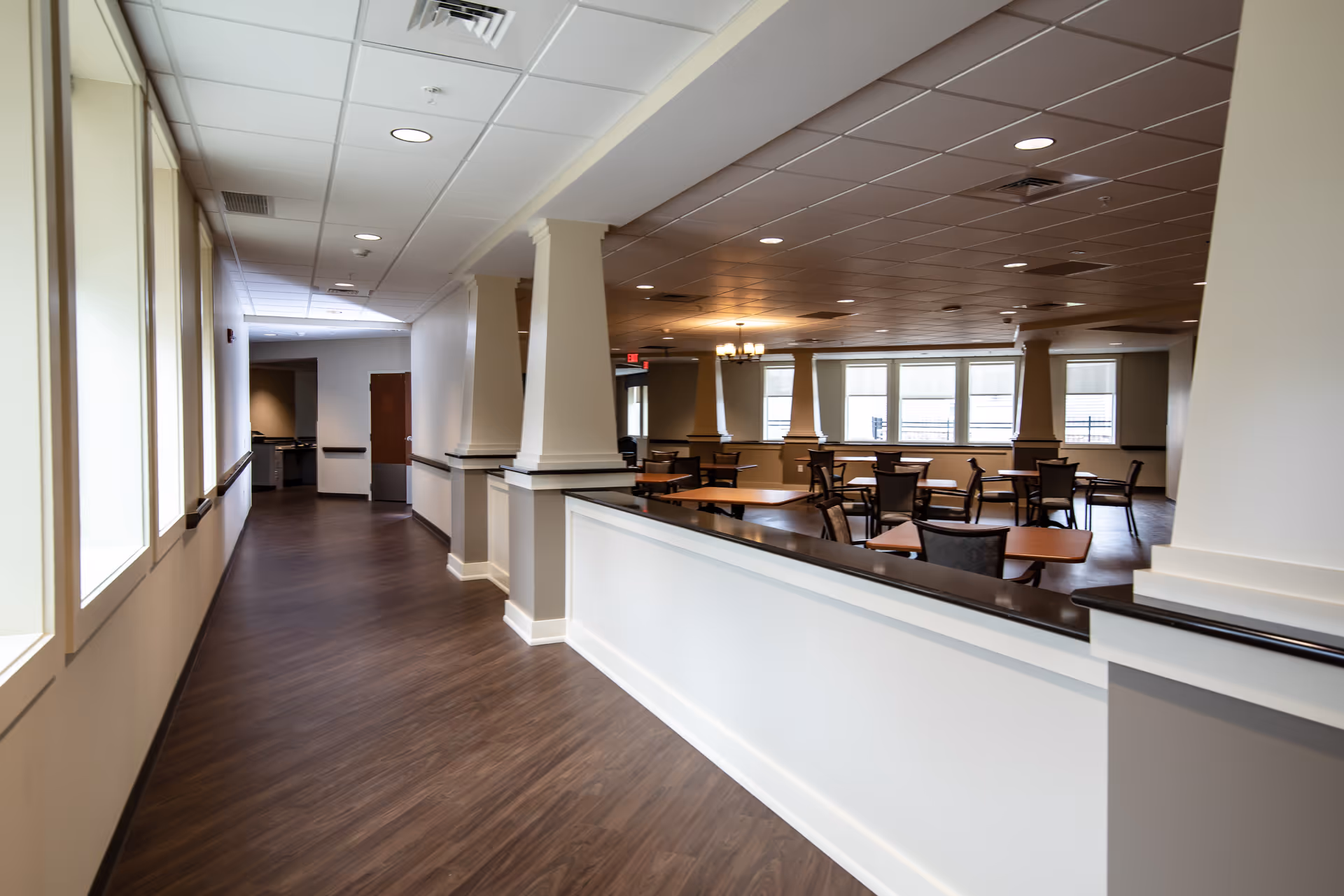 Interior view of a senior living facility hallway leading to a dining area with multiple tables and chairs. The space features wood flooring, large windows, white walls with beige accents, and ceiling lights.