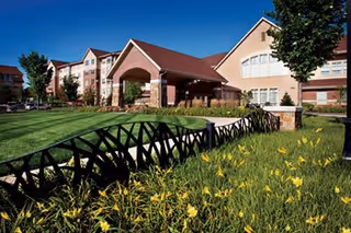 Front exterior of a senior living community featuring the building entrance, manicured lawn, yellow flowers, and a clear blue sky.