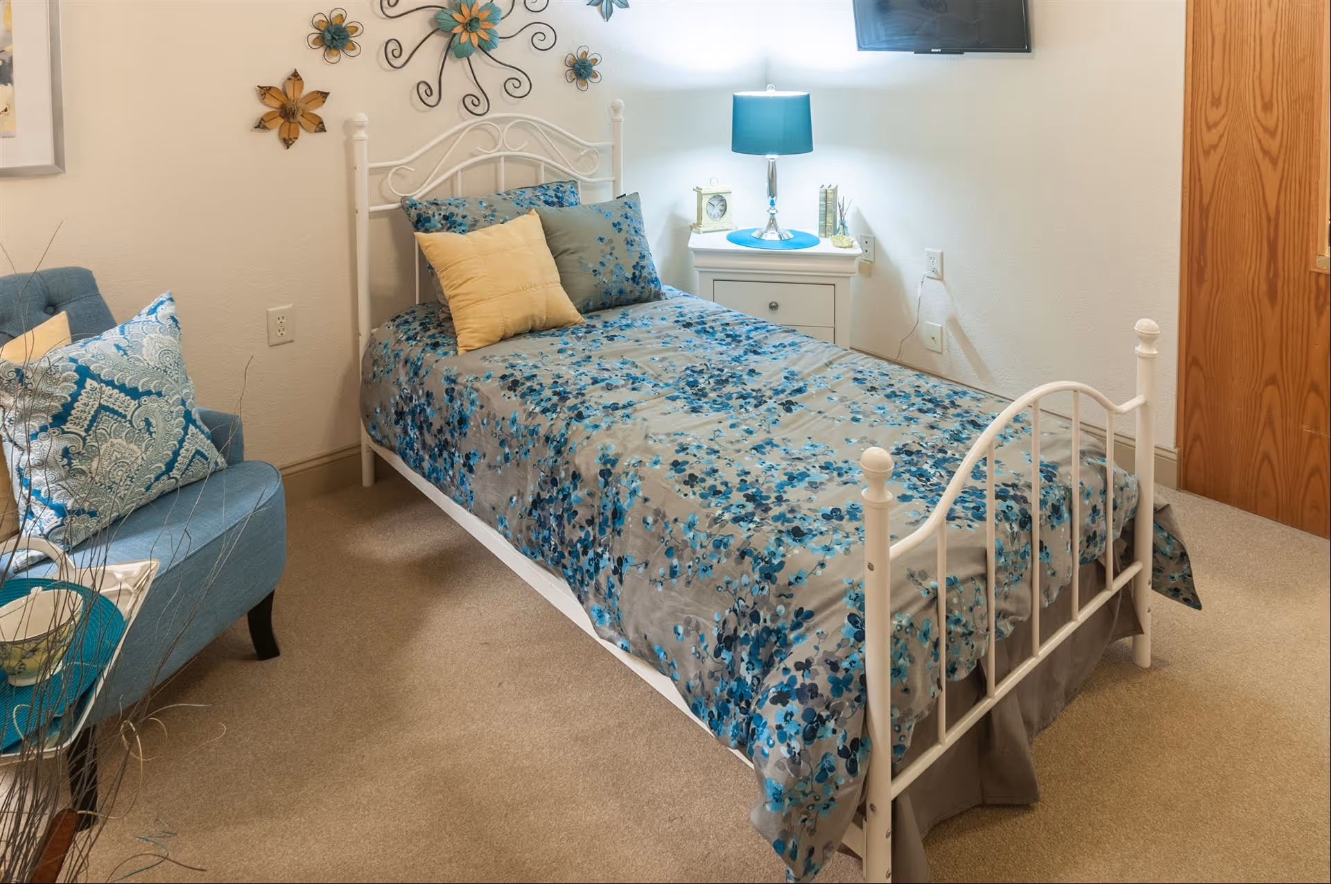 A cozy bedroom with a single white metal bed frame, a bedspread with blue floral patterns, and several pillows including a yellow one. Next to the bed is a white nightstand with a blue lamp, a clock, and some decorative items. On the wall above the bed are metal flower wall decorations. A blue armchair with patterned pillows is positioned to the left, and a wooden door is visible on the right side of the room.