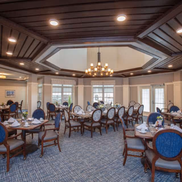 A spacious dining room with multiple round tables set with white napkins, plates, and glassware. The room features blue upholstered chairs with wooden frames, large windows allowing natural light, and a decorative chandelier hanging from a coffered ceiling with recessed lighting.